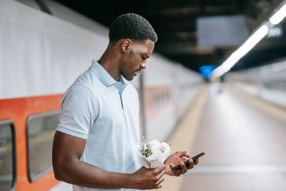 A young man with dark skin and short curly hair, dressed in a light blue polo shirt, is standing on an underground train platform holding a small bouquet of fresh white flowers wrapped in white paper.
