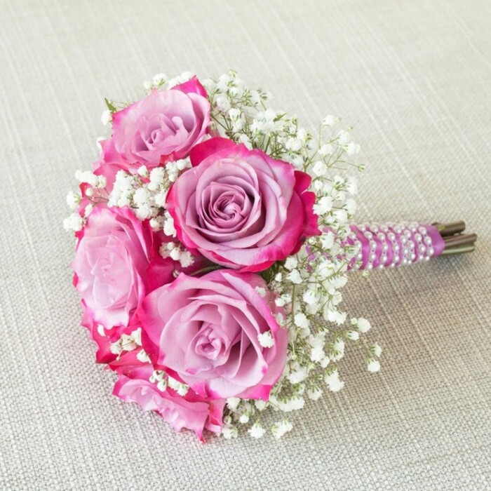 Laid gently on an off-white, linen-like fabric, this exquisite Oxford bridal bouquet radiates artistry and romance. Five sumptuous roses, resplendent in their colour play, draw the eye with petals that blend lavender and pale mauve at their centres, gently dissolving into vivid magenta and rich fuchsia along the outer edges. The shape is rounded and compact, perfect for a bride or bridesmaid wishing to carry something that embodies joy and understated grandeur. Tiny, white baby's breath weaves in and out, imparting a light, airy quality that contrasts with the roses' velvet softness. The meticulously gathered stems are wrapped in a regal, dark purple satin ribbon, adorned by a series of white pearls that spiral gracefully, reminiscent of Oxford's time-honoured traditions. Touches of natural green and brown peek through at the base, reinforcing the fresh-picked authenticity locals expect from Flowers Oxford. The interplay of shades and delicate textures is highlighted by gentle daylight, with the bouquet poised for an intimate wedding at Magdalen College or a romantic gathering overlooking the Isis. Each element invites admiration, making it ideal for local celebrations where elegance and contemporary style are always in bloom.