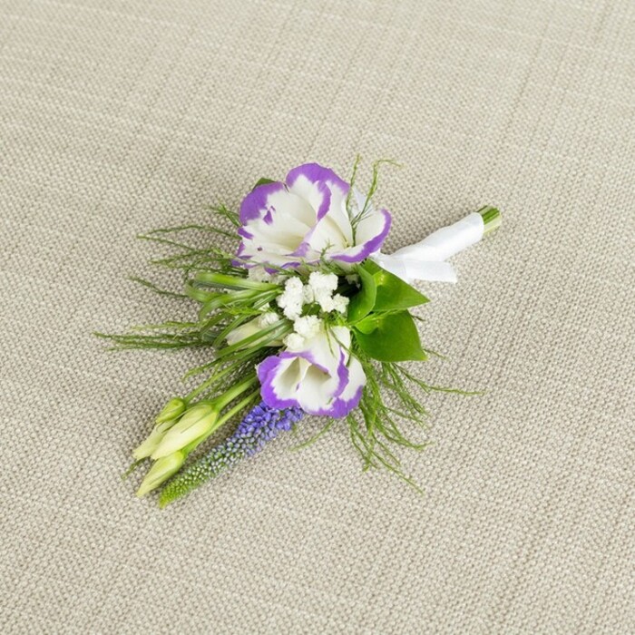 A carefully composed wedding boutonniere is laid out on a soft, light beige linen surface, the faint weave of the fabric adding quiet texture while keeping the focus firmly on the flowers themselves. At the centre, two lisianthus blooms unfold in full detail, their white petals edged with an intense, royal purple that ruffles along each margin, giving the flowers a sculpted, almost watercolor effect. Around these focal blooms, clusters of tiny pristine white blossoms-reminiscent of baby's breath-fill the gaps with a misty, romantic lightness, making the design feel full yet never heavy. Standing upright through the composition, a slim spike of muscari, or grape hyacinth, displays its tight row of deep purple, bell-shaped flowers, adding a vertical line and a richer shade that grounds the palette. Two unopened, pale green buds nestle close by, hinting at new blooms to come and evoking that sense of new beginnings so central to weddings and milestone celebrations in Oxford's historic churches and registry offices. Fine, feathery greenery, similar to asparagus fern, fans out from the base, its delicate fronds softening the edges and lending a natural, garden-picked charm while still appearing crafted and intentional. All the stems are gathered together at the base and wrapped in a clean band of white satin ribbon, tied with a small, elegant bow that indicates the boutonniere is finished and ready to pin onto a lapel. The light appears gentle and even, enhancing the contrast between the cool purples, crisp whites, and fresh greens. Created with the eye of an experienced local florist, this boutonniere would suit a groom or groomsman walking through the cobbled streets of Oxford on the way to an intimate ceremony or a formal evening celebration.