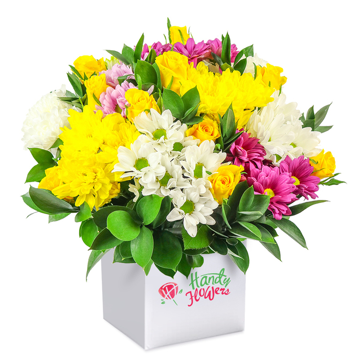 A cheerful, dome-shaped bouquet overflows from a pristine white square box, placed against a seamless white background that allows the lively colours and textures to take centre stage. The composition is dense and abundant, recalling the layered borders of a well-loved Oxford garden in Headington at the height of spring. Dominating the display are sunny yellow flowers: clusters of small yellow roses, some tightly furled and others starting to reveal their inner petals, sit alongside large, plush yellow chrysanthemums. Their softly ruffled surfaces and rounded forms create a sense of warmth and generosity, like sunlight collected in petals. Interwoven among these are cool white blooms, including daisy-like chrysanthemums with neat green eyes and fuller white mums, adding crisp clarity and visual breathing space. Running through the yellow and white are vivid fuchsia and rich magenta chrysanthemums, some with hints of softer pink at the tips, their layered petals adding depth and a touch of drama, similar to the bold colour accents you might see around Oxford's Botanic Garden beds. Around and beneath the flowers, glossy, dark green foliage forms a supportive base, its firm, elongated leaves providing contrast and making the colours appear even more radiant. The white cube box has clean, straight edges and a smooth finish, acting as a minimalist frame for the rounded bouquet and giving the whole piece a contemporary, gift-ready look. This arrangement feels ideal for an affordable yet impressive birthday surprise, a bright get-well gesture to a friend staying near the John Radcliffe Hospital, or a simple way to bring a burst of sunshine and fresh fragrance into a home in East Oxford.