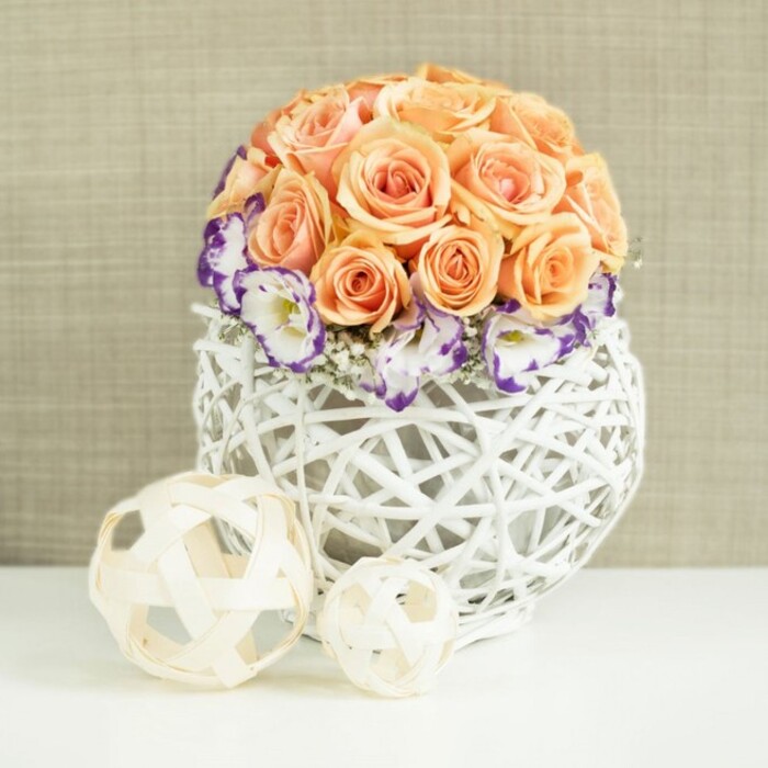 On a clean, white tabletop that could easily belong in a light-filled Oxford townhouse near North Parade, a graceful floral centerpiece takes centre stage. The eye is immediately drawn to a lush, hemispherical mound of roses, arranged so precisely that their soft peach and apricot tones blend into one continuous sweep of colour. Each rose is fully formed, with layered petals that appear almost satin-like, creating a velvety texture across the dome. Just below this crown of roses, a ring of lisianthus blooms gently fans out. These flowers are predominantly white, yet each ruffled petal is finely edged with deep purple or violet, giving an impression of hand-painted detail and adding a sophisticated contrast to the warm peach hues above. Scattered among the lisianthus, airy sprigs of white gypsophila peek through, their tiny blossoms catching the light and lending a misty softness to the base of the arrangement. All of this floral abundance rises from a distinctive round container crafted from white wicker or rattan, its strands woven into a bulbous, open lattice that resembles a delicate basket or sculptural nest. The container's bright white tone echoes the tabletop and amplifies the freshness of the design. In front of the main arrangement, two decorative spheres rest casually: both formed from pale, natural wood or bamboo strips, one noticeably larger than the other, each sphere echoing the same open-weave texture as the vase. Behind the scene, a softly textured beige fabric backdrop, suggestive of fine linen found in Oxford boutiques, lends a warm, neutral setting that allows the flowers to glow. The overall composition feels romantic, serene, and meticulously crafted, perfect for illustrating wedding table decor, refined home styling, or special occasion flowers delivered throughout Oxford and its surrounding neighbourhoods.