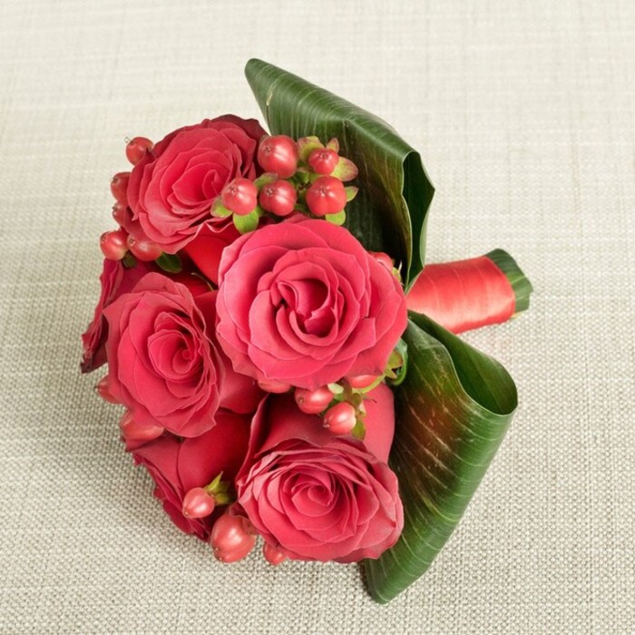 A compact, romantic bouquet of deep red roses and berries lies against a pale, finely textured beige fabric, like a cherished arrangement set down on a table in a quiet Oxford townhouse. Viewed from above at a slight angle, the bouquet appears deliberately rounded and carefully composed. At its centre are several fully opened red roses, their petals thick and velvety, spiralling outward in overlapping layers that range from dark, almost wine-red in the depths to brighter crimson near the edges. Nestled among these blooms, clusters of small Hypericum berries in reddish-pink and muted cherry tones provide delightful dots of texture, each smooth sphere catching the light differently and breaking up the sea of petals. Encircling the base, broad, deep green leaves are elegantly curled and wrapped, forming a sleek, architectural frame that gently lifts and supports the flowers. The leaves' glossy finish and visible veins add a modern, tropical touch, contrasting beautifully with the classic romance of the roses. A slim red satin ribbon is wrapped snugly around the stems, glimpsed near the side of the bouquet, its subtle sheen reinforcing the sense of care and craftsmanship behind the design. The lighting is soft and even, with no stark shadows, allowing the viewer to appreciate the interplay of textures-from the plush petals to the smooth berries and the slightly coarse fabric background. This arrangement feels perfectly suited for a Valentine's delivery to a flat near Oxford Castle, a proposal in University Parks, or an anniversary dinner at a restaurant off Cowley Road, bringing with it the quiet promise of affection and a touch of luxury to everyday moments.