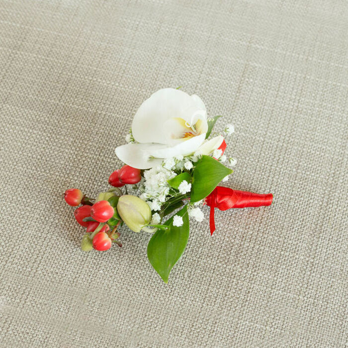 A single, refined floral boutonniere is presented against a neutral backdrop of light beige, woven linen, the subtle texture of the fabric adding an organic, slightly rustic note that enhances the natural beauty of the flowers. At the very centre of the arrangement is a flawless white Phalaenopsis orchid, its broad, gently arched petals unfolding gracefully like soft porcelain wings. The orchid's throat glows with a muted blend of creamy yellow and soft orange, providing a gentle warmth that draws the eye inward. Surrounding this central bloom are delicate clusters of white baby's breath, each tiny gypsophila flower forming part of a light, airy halo that softens the boutonniere's outline and adds fine, lacy texture. Tucked to one side is a small but vivid cluster of glossy red berries, reminiscent of hypericum, their rounded shapes and deep, festive crimson tones suggesting a winter celebration or holiday wedding in Oxford's historic city centre. A few pale green berry buds mingle with them, adding nuance and a sense of natural progression from bud to ripe fruit. Various shades of rich green foliage are woven through the design, with some leaves broad and pointed, others narrower and elongated, framing the orchid and berries with a fresh, verdant border. At the base, the stems are firmly bound with a bright red satin ribbon, its surface sleek and gently reflective, tied into a compact knot with short, tidy tails that rest elegantly on the fabric. The entire composition feels both modern and timeless, ideal for a groom's lapel, a prom guest, or a formal dinner near the Oxford colleges. Carefully composed, it reflects the kind of attentive, detail-driven floristry that clients across Oxford, from Summertown to Cowley Road, look for when marking meaningful occasions.