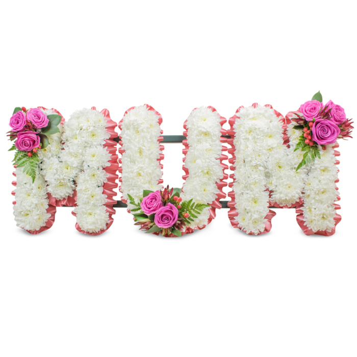 A solemn yet tender standing tribute that reads MUM in large block letters, each letter carefully formed from a mass of small, pure white chrysanthemums. The densely packed mums create a plush, even texture that gives the letters a serene, cloud-like presence against a plain white background. The outside edges of each character are trimmed with a soft ruffled ribbon in pale pink, a detail that brings a gentle warmth and motherly softness to the otherwise crisp composition; a dark green support frame is faintly visible between letters, hinting at the craftsmanship and structure keeping the tribute upright. Three focal accent clusters punctuate the arrangement: the left shoulder of the first 'M' is adorned with a tight posy of vivid fuchsia-pink roses, ferny greenery and tiny red-orange hypericum berries; the 'U' carries the same mix tucked into its lower curve; and the final 'M' is balanced by an identical cluster at its upper right. The contrast between the cool, dignified chrysanthemums and the rich, velvety roses creates a comforting visual rhythm, while the berries and glossy foliage add liveliness and texture. In soft indoor light - the sort that filters through stained glass or a chapel window near the Oxford Covered Market or Broad Street - the tribute feels quietly elegant and profoundly respectful. This composition is conceived specifically for memorials, funerals, and graveside services where families seek a tasteful and personal expression of love and remembrance for their mother.