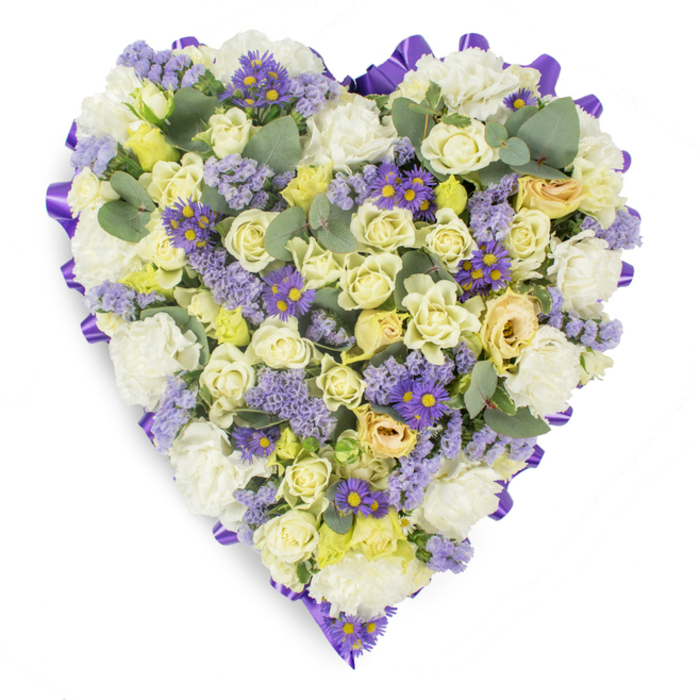 A meticulously arranged heart-shaped floral tribute, shown on a stark white backdrop to emphasize its delicate colours and careful construction. The body of the heart is densely filled with cream and pale lemon roses, their varied openness lending depth and a luxurious, tactile quality; among them, clusters of pristine white carnations build a soft, voluminous foundation. Interspersed throughout are sprays of lavender statice and cheerful deep-purple, aster-like blooms with bright yellow centres, which introduce textural contrast and small, bright accents that animate the composition. Blue-green eucalyptus leaves-round and softly glaucous-are placed at intervals to introduce calm, cooling tones and to relieve the density of the flower heads with graceful, airy shapes. A continuous purple satin ribbon, folded into neat loops around the perimeter, frames the heart and provides an elegant, formal edge suitable for a funeral chapel or a graveside display. The overall palette reads as serene and consoling, the lighting soft and even to capture the gentle gradations between cream, pale yellow, and lavender. This design is intended as a heartfelt sympathy offering: a quiet declaration of love and remembrance, crafted with the precision and care you would expect from a florist serving families across Oxford near Magdalen Bridge and the Covered Market. It conveys respect, peacefulness, and the enduring memory of a cherished life in a manner both classic and tender.
