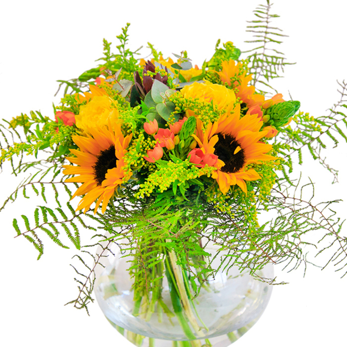 Bright and beaming, this floral arrangement captures an Oxford summer at its heartiest, gathering the city's natural brilliance into one generous bouquet. Sequenced within a flawless, round glass vase, golden sunflowers and layers of buttery-yellow garden roses radiate warmth, each petal unfurling with joyful intent. Their luminous faces are offset by lustrous green foliage-trailing ferns, delicate goldenrod, slender spikes, and the tranquil blue-green hint of eucalyptus-all artfully layered by experienced hands. Glossy red Hypericum berries peek from between the flowers and greens, their jewel tones glistening like berries found in the wilder hedgerows along Port Meadow. The transparent vessel allows a close view of the stems, immersed in water, their lines forming a living architecture reminiscent of the elegant bridges crossing Oxford's rivers. Up close, the textural contrast between smooth petals, plush roses, suede-like sunflowers, and the crisp snap of fresh greenery invites a multi-sensory experience, heightened by a light, invigorating scent. The bouquet's exuberance makes it a wonderful companion for moments of celebration: a birthday in Jericho, a summer dinner in Summertown, a new beginning for an Oxford student. Each element is thoughtfully chosen, infusing the arrangement with the warmth and hospitality for which the city is known.