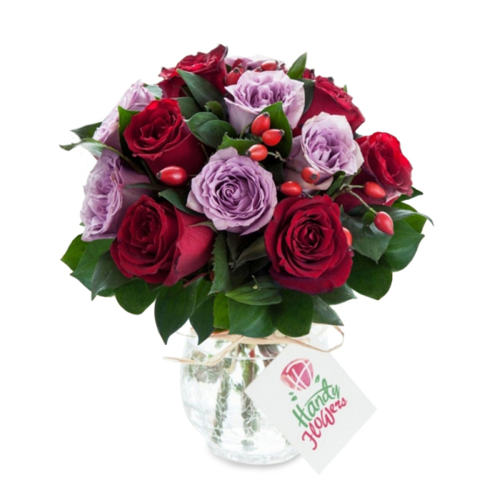 A sumptuous, intimate arrangement blending deep crimson and burgundy roses with soft lilac and dusty purple roses, gathered into a dense, rounded silhouette that feels both luxurious and approachable. Each rose appears newly opened, the petals layered and slightly translucent at the edges; between them, lush dark green foliage-probably Salal or Ruscus-adds cool, structured contrast and helps the colours read more vivid. Dotted throughout are bright red-orange rose hips, their small, glossy forms bringing an organic, almost hedgerow-like texture that complements the roses' softness. The bouquet sits in a decorative mercury-effect glass vase: an antique-silver, crackled surface that reflects highlights and lends a slightly vintage elegance. The vase's bulbous body tapers toward the neck, where a natural raffia tie is knotted and a white rectangular gift tag tilts forward bearing a small rose illustration and space for a handwritten sentiment. Set against a clean white background, the arrangement's tones pop and the metallic vase shimmers; you can almost sense the cool satin of the petals, the faint green scent of foliage, and the tactile contrast of raffia and glass. As a florist serving Oxford - from Summertown down to Headington and the lanes around University Parks - I imagine this bouquet arriving at a Georgian townhouse, a college celebration, or as a thoughtful anniversary gift. Crafted with local sensibility by Flowers Oxford, it reads as a carefully made, romantic choice for marking special days or offering quiet reassurance to someone you love.