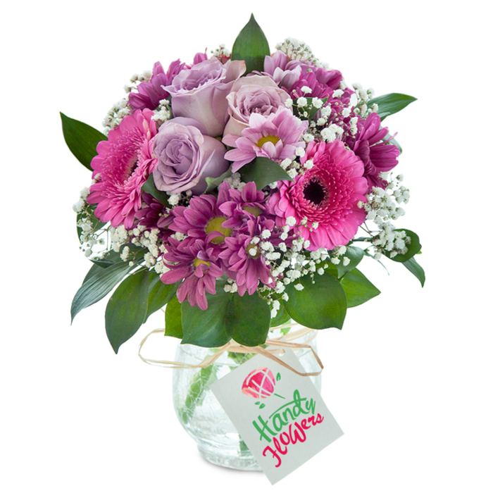 This lavish, handcrafted bouquet, arranged by a professional Oxford florist, radiates vibrant energy with every detail. Central among the plush blooms are roses in soft lavender shades, their petals unfolding with grace, symbolizing heartfelt wishes akin to those exchanged beneath Oxford's dreaming spires. Bold fuchsia Gerbera daisies provide striking contrast, their large faces and inky centres catching the eye much like the lively storefronts of Summertown. Encircling the focal blooms are velvety magenta chrysanthemums, each petal adding dimension and a subtle play of colour. Delicate white gypsophila is interspersed, creating a gentle, airy feel reminiscent of the crisp, clean air floating over the lawns of University Parks. Deep green leaves build a lush foundation, grounding the arrangement and echoing the old trees of Headington Hill. The bouquet is housed in a distinctive clear glass vase with an attractive crackled texture, adding depth and sparkle under natural light, and is tied at the neck with a simple raffia ribbon that hints at rustic charm. A neat, white rectangular tag completes the design, ready to hold a personal message for a special Oxford occasion-be it a graduation, a birthday, or a thoughtful thank you. Crafted with a deep understanding of local tastes and traditions, this bouquet offers a visual and aromatic celebration of Oxford's refined, welcoming spirit.