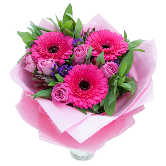 I often think the charm of pink flowers lies in their ability to feel both playful and gracious, a quality on full display here in this hand-tied arrangement. Three large gerbera daisies, vivid in fuchsia-pink, open confidently with sculptural layers of petals surrounding deep, moody centres-their bold colour immediately draws the eye. Nestled among them are several roses in a softer, dusty lavender-pink, their petals tightly curled like the promise of a soon-to-open bloom and offering a velvety counterpoint to the gerberas' graphic shapes. Sprays of small purple statice tuck into the gaps, providing a speckled texture and a cool violet note that punctuates the warm pinks. The bouquet rests against a generous bed of green foliage-broad leaves that might be salal or eucalyptus-whose crisp sheen and subtle fragrance add an almost garden-fresh aura. The florist's wrapping is part of the composition: an inner layer of pale pink, matte tissue cushions the stems while a layer of translucent fuchsia-pink cellophane, slightly crinkled, catches and softens the light, unifying the colour story and lending a celebratory sheen. You can see the top rim of a clear glass vase at the base, a small reveal that suggests immediate display on a sideboard or coffee table. The overall mood is intimate and buoyant, perfect for marking birthdays, Mother's Day, anniversaries or simply bringing a burst of optimism. As someone who arranges flowers for neighbours across Cowley and Headington, I picture this bouquet greeting a loved one after a stroll through the Botanic Garden or brightening a flat near the Oxford Covered Market.