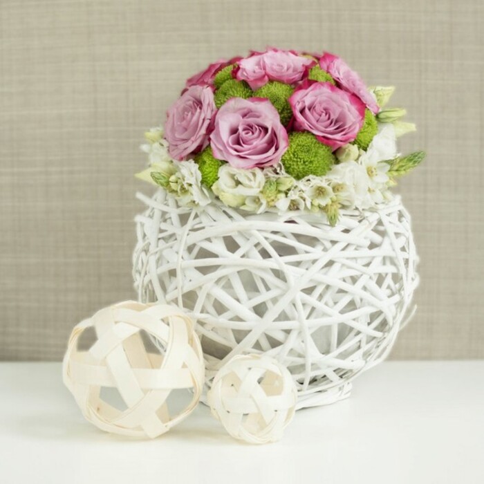 A romantic, dome-shaped bouquet brimming with pastel blooms is shown resting in a distinctive white woven vase, placed on a smooth white surface against a softly blurred beige backdrop that recalls the calm interiors of an Oxford townhouse. The vase itself is spherical and made from interlaced white branches or twig-like strands, creating an open, airy lattice through which shadows gently fall, giving a sense of depth and lightness. From this sculptural base rises a compact arrangement of roses in a harmony of soft pink and gentle lavender shades, each bloom fully open so that layers of velvety petals swirl toward the centre. Some petals are edged with slightly deeper pink, adding definition and a hint of drama without disturbing the overall softness. Nestled among the roses are bright green button chrysanthemums, their small, rounded heads offering a tactile, almost mossy texture and a cheerful pop of colour. Around the lower edge of the bouquet, dainty white and cream filler flowers thread their way between the larger blooms, forming a delicate collar that helps transition from flowers to vase. To the left of the main arrangement, two pale wooden or rattan spheres rest on the tabletop, one larger, one smaller, each with an open, woven design that echoes the vase and introduces a natural, handcrafted note. The neutral beige background, reminiscent of fine linen or burlap, keeps attention on the flowers while adding warmth and subtle texture. This serene, beautifully balanced arrangement would sit perfectly on a wedding table at an Oxford college dining hall or as an elegant centrepiece for an intimate celebration along the banks of the Cherwell.