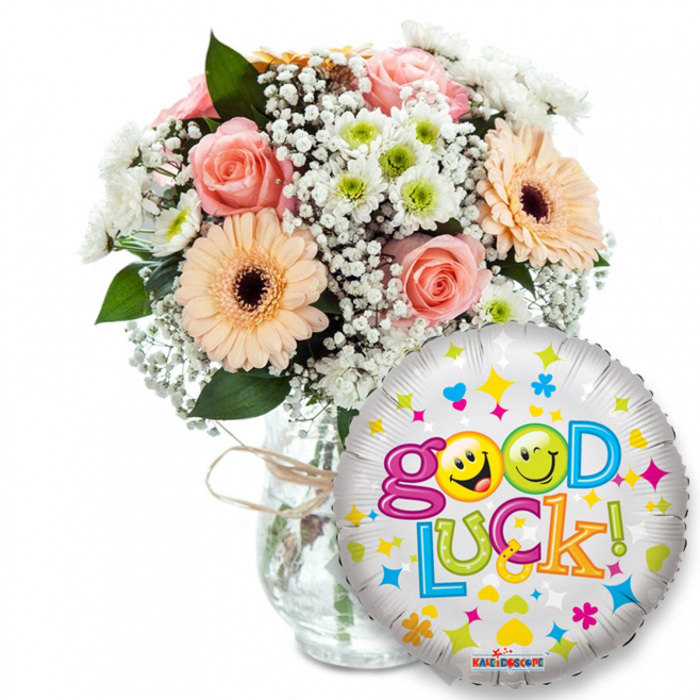 Set against a pristine white background, this supportive 'Good Luck!' gift reads like a small celebration captured in still life. On the left a generous bouquet rests in a clear glass vase wrapped with a simple twine tie; the florist's hand is evident in the relaxed, abundant styling. The floral mix pairs soft peach and pale pink roses with radiant peach-orange Gerbera daisies - the daisies' dark brown centres grounding the composition - while clouds of tiny white baby's breath weave delicately between blooms. Clusters of tiny white and lime-green chrysanthemum-style florets add crisp, cheerful texture and a subtle, clean scent that contrasts with the roses' soft perfume. Glossy deep green foliage peeks through, accentuating the pastel tones and giving the arrangement healthy structure.In the foreground to the right, a round silver foil balloon overlaps the bouquet and introduces a celebratory graphic note. Its metallic surface sparkles and is strewn with multicoloured confetti-like shapes - stars, hearts and small cross motifs in pinks, blues, yellows and greens. The phrase Good Luck! appears in a joyful, varied palette of letters, and playful yellow smiley faces are incorporated into the typography, lending the balloon a warm, human touch. The balloon's lower edge bears a small KALEIDOSCOPE logo, a discreet designer mark.The overall impression is hopeful, buoyant and reassuring - the kind of gift one might have delivered across Oxford from the High Street to Magdalen Bridge for a graduation, a farewell tea, or the morning of an important interview. The tactile contrasts - satiny rose petals, papery Gerbera petals, feathery Gypsophila and the smooth sheen of Mylar - make it a sensory embrace as well as a visual one.