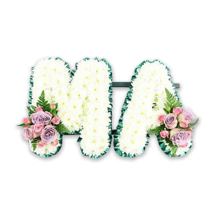 This photograph features a personalised funeral letter tribute spelling MA, designed as a graceful homage to a much-loved mother or maternal figure. Both letters, shown in bold capitals, are completely enrobed in small white chrysanthemum blooms, each floret carefully placed so the surface appears even, full, and luxuriously padded. The white petals, slightly ruffled at the edges, catch the soft light, giving the tribute a gentle glow against the plain white background. A dark emerald green border follows the outline of the M and the A, likely formed from pleated satin ribbon or a neat band of foliage, which crisply defines the shapes and provides a sophisticated contrast to the whiteness within. At the lower left foot of the M and the lower right foot of the A, harmonising flower sprays are arranged as tender accents. Here, pale pink roses, dusky lilac roses, and petite pink spray roses come together in a delicate blend of pastel tones. Some heads are fully open, revealing their spiralled centres, while others remain in bud, suggesting continuing love and memory. Fine green fern fronds and small leaves weave through the roses, creating depth and soft texture. The overall layout is balanced and symmetrical, providing a sense of calm order and reverence. This kind of floral lettering is often requested by families in Oxford for services at local churches or crematoria, a tangible way to write a cherished name or relationship in flowers. The tribute in the image conveys deep affection and respect, providing a comforting focal point beside the coffin or at the front of the chapel during a final farewell.