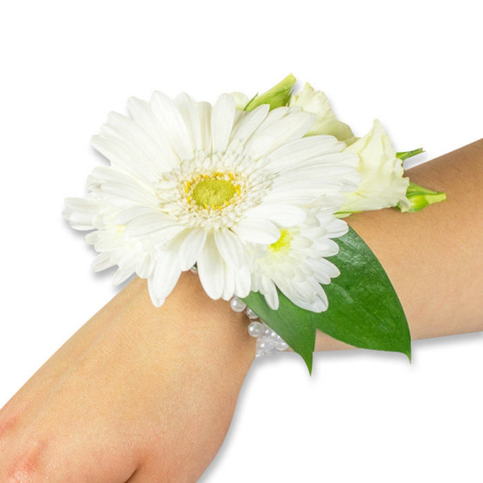 A striking white floral wrist corsage is photographed on a fair to medium forearm against a crisp white background, its refined silhouette reflecting classic bridal sensibility with a touch of local character. The focal Gerbera, a large white bloom with abundant, velvety petals, radiates from a bright lime-green and soft yellow center that adds a subtle modern accent to the otherwise timeless palette. Nestled close to this central blossom are smaller pure-white cluster flowers-resembling spray chrysanthemums-and a handful of creamy lisianthus, some in tight bud and some with petals unfurling, which together create gentle depth and a soft-textured crown. Beneath and between the flowers, deep green salal or Ruscus leaves form a lush backdrop; their broad, glossy surfaces ground the arrangement and bring out the clean clarity of the whites. The whole cluster is fastened to the wrist by a sophisticated, multi-strand band of glimmering faux pearls that hug the skin and reflect light with a whisper of sheen, lending old-world charm to a contemporary design. Imagining the piece in daylight-perhaps on High Street as sunlight filters through college gates-you would notice the contrast between cool, smooth petals and the taut resilience of the foliage, and sense the delicate, fresh scent of cut stems. Suitable for weddings, proms, and milestone ceremonies, this corsage reads as both an elegant accessory and a small, wearable memory, thoughtfully composed by a florist who understands proportion, occasion, and the quiet beauty of Oxford moments.