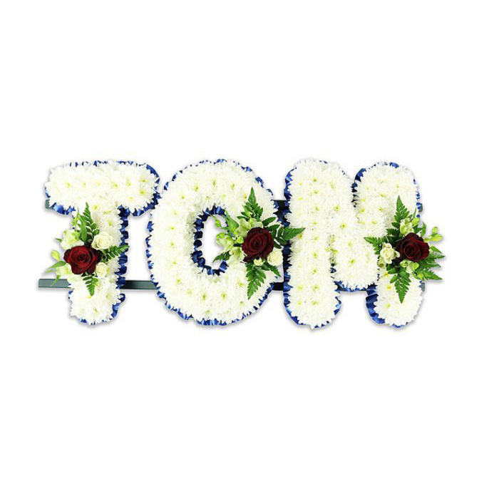 A formal, personalised floral memorial that spells TOM in large, sculpted letters, each letter densely covered with countless small white chrysanthemum-like blooms to create a plush, three-dimensional surface. The white chrysanthemums are arranged tightly so the letters hold a soft, cloud-like volume, while a precise royal blue ribbon neatly outlines every edge, adding crisp definition and a respectful accent of colour. At the lower or central portion of each letter sits a composed spray: a single deep red rose forms the focal heart of the spray, symbolising love and remembrance, with clusters of small creamy spray roses and delicate fern fronds fanning around it to add visual depth and texture. A subtle dark green or black metal support bar is visible behind the base of the letters, providing stability so the tribute can be displayed upright on easels or stands in chapels and crematoria. The image is set against a stark white background with even, neutral lighting, which keeps the focus on the floral craftsmanship and the interplay of colour-white, royal blue, rich red and fresh green-without distraction. This arrangement is the sort of bespoke bereavement piece frequently requested by families across Oxford, whether for a service near Summertown, a wake by Jericho's community halls, or a remembrance gathering close to the Radcliffe Camera. The tone is composed and compassionate, a carefully made floral statement intended to honour a life with clarity and grace.