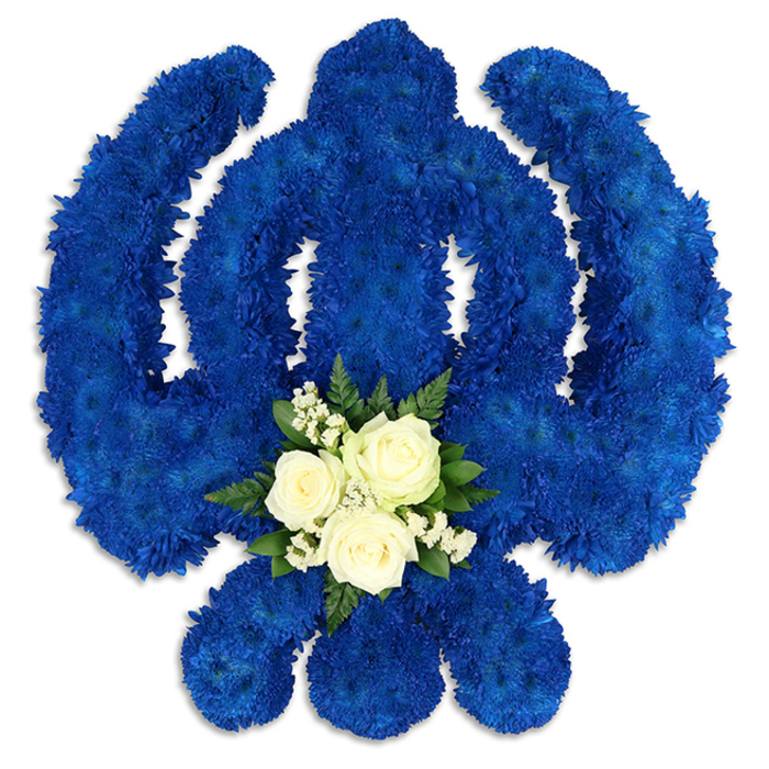 An impressive Sikh Khanda floral tribute fills the frame, presented against a simple white background so that its form and colour speak clearly. The entire symbol-the unbroken circular chakkar, the central upright double-edged sword, and the two elegant, outward-sweeping kirpans-is sculpted from masses of deep royal blue chrysanthemum florets or similar compact blossoms. Their petals overlap to create a plush, almost quilted surface that feels rich and substantial, much like a ceremonial emblem carefully laid out for a significant Sikh funeral or remembrance event in Oxford. The blue tone is intense yet calming, evoking dignity, courage, and spiritual depth. At the base of the central sword, resting where the eye naturally comes to pause, a small contrasting arrangement softens the design. Three pristine white roses, perfectly shaped and just open, form the heart of this cluster. Their creamy petals catch the light gently, suggesting purity, peace, and loving remembrance. Around them, fine sprigs of tiny white filler flowers, likely baby's breath, add delicate texture, while a trim collar of dark green foliage provides balance and a fresh, natural edge. Subtle shadowing beneath the Khanda hints at thickness and craftsmanship, as though the tribute has been carefully built layer by layer on a sturdy base. The overall impression is of a thoughtfully created, culturally resonant piece-something a skilled local florist in Oxford might prepare for a family wishing to honour a relative's Sikh faith at a crematorium service in Botley or a memorial gathering near the city centre, combining solemn respect with quiet beauty.