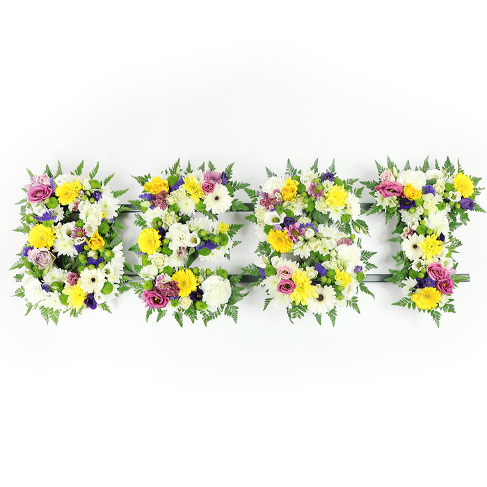 A vibrant, lovingly assembled floral lettering piece reads BEST across a bright white field, each letter formed from a dense, tactile collage of fresh flowers and rich greenery. The dominant tones are cheerful yellow and pristine white: clustered white chrysanthemum-like blooms and cream gerberas create soft, voluminous planes, while yellow roses and gerberas bring warmth and a sunny, comforting energy. Swathes of deep purple lisianthus or similar small blossoms thread quiet, thoughtful shadows through the display, counterbalanced by lively magenta-pink spray roses or carnations that catch the eye like small, affectionate notes. Fine clusters of pale green and white filler blooms add a whisper of texture, and substantial dark-green fern fronds wrap the outer edges of each letter, their feathery tips softening the letterforms and adding a woodland sensibility. A subtle supporting framework in dark green or metal is visible at points - especially across the E's middle bar and lower spans - showing the engineering that keeps this large tribute intact for laying on a plinth or easel. Photographed head-on with even, bright illumination, the piece reads as fresh and handcrafted; you can almost imagine the faint, herbaceous fragrance and the gentle rustle of fern as family and friends gather. This arrangement works beautifully for memorials and remembrances where the sentiment is personal and affirmative - a floral statement that honours a loved one as truly the best.