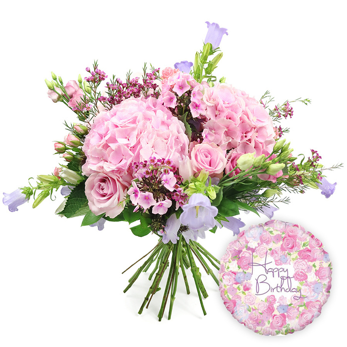 Set against a spotless white backdrop, a hand-tied birthday bouquet fills the frame with soft pinks, purples, and fresh green, like a little pocket of spring lifted straight from an Oxford college garden. The eye is first drawn to several large, rounded heads of blush pink hydrangeas, their petals layered so densely that they appear almost cloud-soft, giving the arrangement a plush, romantic base. Nestled within these voluminous clusters are elegant light pink roses, some fully open to reveal their classic, swirling centres, others still curled in tight buds that hint at the blooms to come. Scattered delicately throughout, sprigs of rich fuchsia-pink waxflowers add sparkle and fine texture, their tiny stars of colour brightening the gentler pastel tones. Rising from within the bouquet, slender campanula stems bear bell-shaped flowers in soothing shades of lavender and pale purple, adding a sense of height and gentle movement as they tilt slightly outward. A mix of verdant foliage frames the flowers: long, tapering leaves and shorter, broader greens weave around the blossoms, creating a structured yet natural outline in a softly domed shape. The gathered stems are clearly visible at the bottom, neatly tied, suggesting this is a freshly crafted bouquet ready to be carried down St Giles' or delivered to a birthday celebration in Headington or Botley. To the lower right, a round Mylar Happy Birthday balloon gleams with a subtle, metallic sheen. Its surface is covered in a pretty, all-over pattern of tiny roses, buds, and foliage in pink, lavender, and cream, mirroring the bouquet's palette. In the centre, graceful purple script spells out Happy Birthday, completing a harmonious and joyful gift ensemble that feels both celebratory and tender.