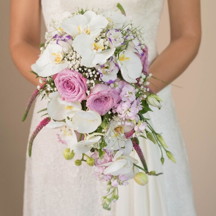 With exceptional care and artistry, this bridal bouquet stands as a testament to Oxford's floral heritage-crafted for a bride whose arms draw close a profusion of blooms that seem plucked from the most enchanting secret gardens off Cowley Road. The star of the arrangement, pure white Phalaenopsis orchids, radiate luxury, their delicate petals arching gracefully and catching light as they cascade amongst clusters of blushing pink and lavender-hued roses. The soft murmur of gypsophila, with its tiny white stars, adds a sense of dreamy movement and a cloud-like softness whispering throughout. Imbued with the subtle fragrance of fresh roses and the gentle spice of amaranthus, the bouquet feels simultaneously modern and timeless. Slender, magenta trails of amaranthus tumble downward, bringing a vibrant pop of colour and distinctive texture, reminiscent of wild botanicals in the meadows beyond Port Meadow. Intermittent green foliage and unopened flower buds peek through, enriching the natural effect and gently connecting the bouquet to the fresh air of Oxford's many parks. The bride's lace gown, luminous and understated, provides the perfect backdrop for the vibrant florals to shine. For those planning a ceremony in Oxford, this bouquet is more than just a floral accent-it's a poetic expression of love, tradition, and new beginnings inspired by the city's unique charm.