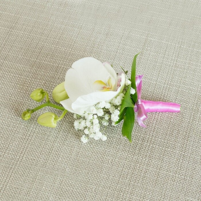 On a softly woven, light oatmeal-coloured fabric that recalls a linen napkin from a small Oxford café, a refined floral boutonniere or corsage is displayed horizontally, inviting the eye to travel along its length. At the centre of this carefully crafted piece is a pristine white orchid, likely a Phalaenopsis, with broad, smooth petals that open in a gentle, symmetrical curve. The orchid's heart reveals subtle touches of soft yellow and a whisper of pale pink, giving depth and character to the otherwise pure white bloom. Nestled at its base, numerous tiny blossoms of white baby's breath gather in airy clusters, each miniature flower adding to a cloud-like, romantic texture that softens the overall silhouette. A few slender, bright green leaves emerge from beneath and behind the flowers, introducing a lively contrast that makes the white petals seem even more luminous. Extending toward one side, a short stem holds a pair of tightly closed, pale green orchid buds, their rounded shapes hinting at future blossoms and giving the arrangement a sense of natural growth and movement. The stems are impeccably gathered and wrapped with a vivid fuchsia pink satin ribbon, tied into a small, elegant bow with short, neat tails that lie smoothly against the fabric. The slight sheen of the ribbon offers a joyful accent of colour that suggests celebration without overwhelming the calm palette. Gentle, even lighting reveals the soft textures of the petals, the intricate structure of the Gypsophila, and the subtle weave of the background. This classic design feels ideal for a groom's lapel at an Oxford wedding near Christ Church Meadow or a formal event at one of the colleges, capturing a polished yet tender elegance. Crafted in the style favoured by experienced local florists, it quietly reflects care, tradition, and attention to detail.