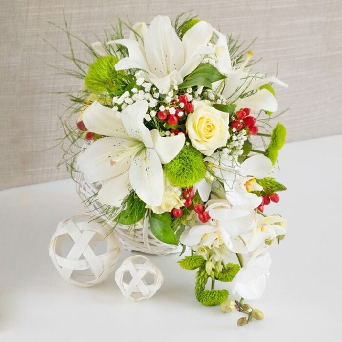 A sophisticated, cascading floral display in tones of white, cream, and fresh green is presented as a statement table arrangement, reminiscent of the elegant celebrations held across central Oxford, from Summertown to the colleges near Christ Church Meadow. The flowers are carefully composed in a round white woven wicker basket, whose subtle texture feels both contemporary and rustic, resting on a smooth white surface that reflects the shapes of the blooms. Several magnificent white Oriental-style lilies form the core of the design, their broad, velvety petals unfurling to reveal speckled throats and prominent stamens dusted with warm pollen. Nestled close to the top, a single creamy ivory rose, perfectly formed, adds a soft golden glow against the cool whites. From the right side, a flowing cascade of white phalaenopsis orchids trails downward, each arching stem dotted with pristine, butterfly-like blooms that create a graceful waterfall effect ideal for weddings and formal events. Around these focal flowers, clusters of bright green pom-pom chrysanthemums introduce a playful, rounded texture that contrasts with the lilies' strong lines, while airy, feathery foliage and sleek dark green leaves build a lush, layered backdrop. Tiny white baby's breath flowers thread through the arrangement like delicate lace, and glossy, deep red berries add vivid pops of colour that feel festive yet refined. To the left of the basket, two decorative white wicker spheres of differing sizes lie on the tabletop, echoing the basket's weave and framing the composition. The soft beige fabric background provides gentle warmth without distraction, allowing the flowers to shine. The overall impression is of expertly crafted, fresh wedding flowers lovingly prepared by an experienced Oxford florist, perfect for a top table, ceremony display, or sophisticated home décor.