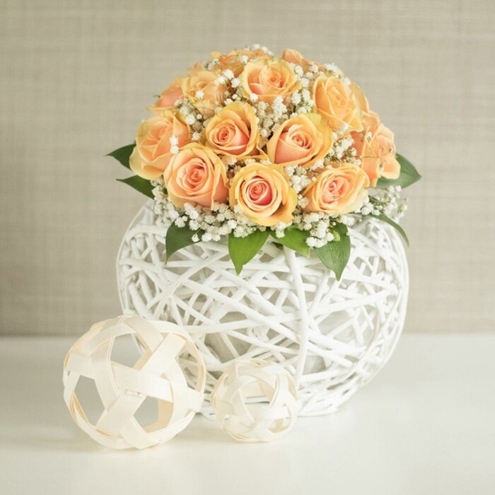 A sophisticated floral centerpiece is arranged on a smooth, bright white tabletop, its focus a rounded bouquet of soft peach and apricot roses nestled into a striking white lattice vase. The bouquet appears full and generous, with roughly two dozen roses placed closely together so that their velvety blooms form a gentle dome. Each rose displays a subtle play of colour: the centers glow with warm, deeper orange tones, while the outer petals soften into pale peach and almost creamy blush, giving the arrangement a sunlit, romantic quality reminiscent of an early evening stroll along Oxford's Iffley Road when the sky begins to warm. Threaded through the roses are numerous clusters of delicate white baby's breath, their tiny blossoms forming airy halos that drift between the larger petals. These small flowers lend a cloud-like texture that lightens the overall look and adds intricate detail when viewed up close. Tucked beneath the blooms, glimpses of dark green foliage offer contrast and a sense of natural structure, as though the bouquet has been freshly gathered from a garden just beyond a historic Oxford college wall. The flowers are housed in a distinctive spherical vase crafted from thin white strands, woven into an open, crisscrossing lattice pattern. This design creates a sense of lightness and transparency, allowing soft shadows and highlights to play through the gaps. In front of the vase, resting directly on the sleek white surface, sit two additional decorative orbs made from similar materials. One is slightly larger and more geometrically defined, while the smaller orb has a more irregular, organic weave, echoing the main vase's texture while adding playful variation. The background is a calm, neutral wall in light beige or greige, subtly textured but unobtrusive, helping the warm roses and crisp white accents to stand out clearly. The overall effect is one of calm elegance and contemporary charm, ideal as a wedding table centerpiece at a small Oxford venue near the Ashmolean, a refined anniversary arrangement, or an example of stylish home decor captured in soft, even lighting that flatters every petal and curve.