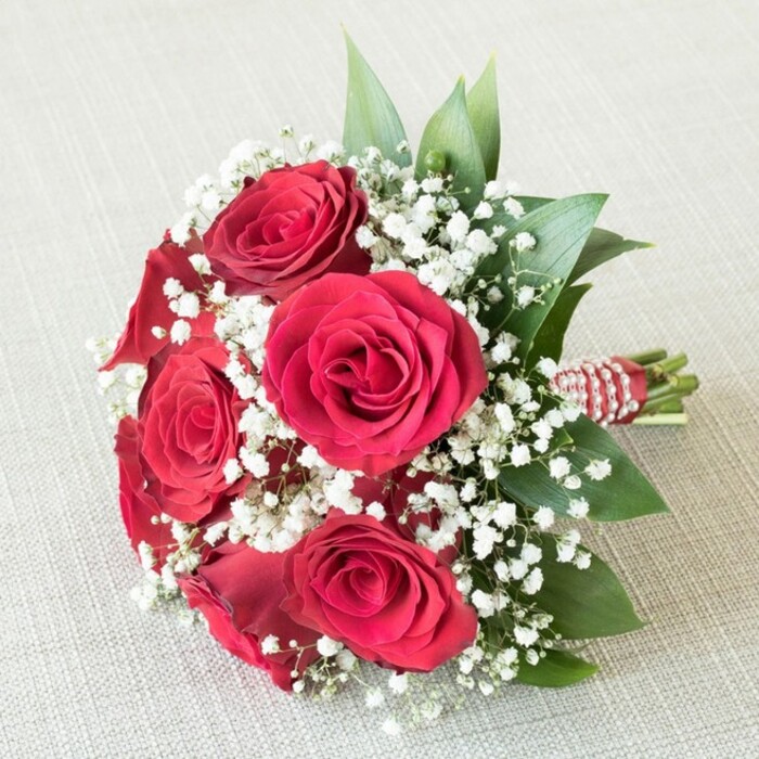 Showcased against a softly textured, light beige background akin to the warm stone facades of Oxford's colleges, this hand-tied bouquet is a masterful study in contrast and harmony. Central to the arrangement are five or six deep crimson red roses, their petals plush and velvety-opening from a tightly swirled center into sculpted, elegant blossoms that speak to the city's poetic heritage. Filling the spaces between the roses are innumerable tiny, cloudlike tufts of pure white Gypsophila, better known as baby's breath, which blanket the bouquet in a gentle, ethereal radiance reminiscent of a misty dawn over Port Meadow. The greenery, likely mature sprigs of salal or ruscus, introduces a fresh, lively green that enhances the depth and framing of the structured bouquet. Each stem is neatly bundled and bound with a strikingly bold red and white striped ribbon, accented by a coil of gleaming white pearls that catch the light, recalling the classic elegance characteristic of Oxford's most cherished celebrations. Every detail, from the natural textures to the interplay of colours, conveys a sense of timeless romance and thoughtful artistry, making this bouquet a fitting centrepiece for weddings, anniversaries, or romantic occasions in Oxford. The even, natural lighting preserves each hue and highlights the impeccable freshness found in every bloom, echoing the expertise and pride of Oxford's finest florists.
