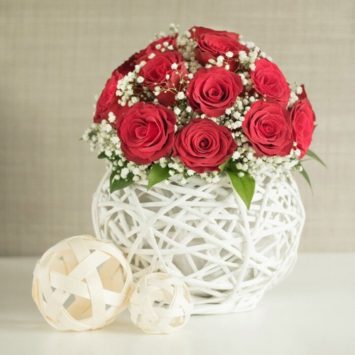 At the heart of this image sits a romantic dome of red roses, composed with the precision of an experienced florist. The blooms are a deep, saturated crimson, each one fully open yet still perfectly shaped, their petals forming tight, spiralling whorls that give the bouquet a plush, almost velour texture. Between the roses, light and airy sprays of white baby's breath weave in and out, the tiny gypsophila blossoms creating a fine, cloud-like veil that both contrasts with and softens the bold red. Here and there, a few dark green leaves emerge, subtle but essential, grounding the arrangement and introducing a natural touch of woodland freshness. This carefully balanced composition is gathered into a rounded, bulbous vase crafted from white interlaced strands. The open-weave design, similar to woven wicker or rattan, wraps around the vase in a loose lattice, allowing the background tone to flicker through the gaps and lending the piece a handmade, artisanal character. The vase stands on a smooth white surface, immaculate and uncluttered, reminiscent of a neatly set wedding table in an Oxford venue near the Bodleian or along St Aldate's. To the left of the main display, two decorative lattice spheres in soft, off-white hues sit casually against the tabletop, their woven strips arranged in a more open pattern. One orb is noticeably larger, with a smaller twin easing back into the mid-ground, giving a sense of dimension and informal elegance. Behind everything, a softly textured wall in pale beige remains out of focus, offering a calm backdrop that allows the rich colours and tactile materials to shine. The overall effect is of a refined, romantic centrepiece, ideal for wedding receptions, anniversary dinners, or intimate gatherings across Oxford.