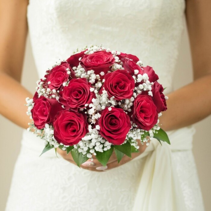 Captured in graceful detail, this chest-level close-up highlights a bride in Oxford holding a superbly crafted round bouquet-a harmonious medley of lush, deep red roses, their petals unfurling in a velvety, tightly woven embrace. Each rose appears at the peak of perfection, and nestled between them are wispy clusters of pure white baby's breath, adding a cloud-like, airy beauty that softens the intensity of the crimson blooms. The bouquet is accented by a scattering of bright green leaves at its base, grounding the design and echoing the natural beauty of Oxford's riverside gardens. The bride's carefully manicured hands support the bouquet with gentle reverence, her fingernails delicately gleaming, while the upper portion of her gown-ornately detailed with lace or embroidery-appears gracefully in the background, its fabric reminiscent of Oxford's sense of scholarly elegance. A sash or length of soft fabric falls fluidly to one side, complementing the overall harmony. The background is gently blurred in cream and beige, focusing attention entirely on the bridal florals and the subtle, tactile details. This bouquet's timeless composition is a floral homage to tradition and romance, ideal for Oxford couples envisioning an elegant celebration or exploring bespoke floral creations for university weddings and chapel ceremonies.
