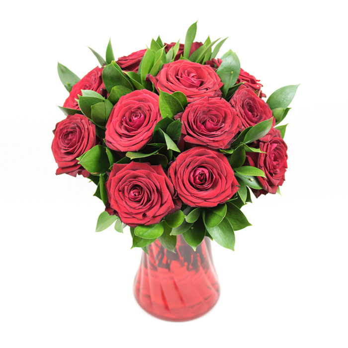In this carefully composed image, a classic round bouquet of about a dozen deep red roses sits in a translucent red glass vase, placed against a pure white background so that every detail is clearly defined. The roses are fully open, each bloom revealing layers of plush, velvety petals that spiral toward the centre in a tight, precise formation, evoking the sense of a perfectly crafted romantic gesture. Their colour is a true, saturated crimson, with slight shifts in tone where the light brushes the edges of the petals, suggesting freshness and quality you might expect from a trusted Oxford florist. Between the blooms, luxuriant green foliage-likely ruscus or a similar leaf-is threaded generously, its smooth, slightly pointed leaves polished to a natural shine. The dark green tones provide a sharp, refreshing contrast to the red, adding depth, roundness, and a touch of garden-like naturalness, reminiscent of the leafy corners of Christ Church Meadow after a rain. The vase is made of red glass with a gentle hourglass curve and a modestly flared neck; a faintly ribbed or swirled texture runs through the glass, catching the light and creating subtle ripples of shadow that give the container dimension without drawing attention away from the flowers. Through the semi-transparent glass, the stems can be seen gathered neatly, grounding the bouquet in a sense of honest simplicity. The clean, even white backdrop allows the arrangement's colours and forms to stand out crisply, making this design feel both modern and timeless-perfect for marking a milestone anniversary, a romantic birthday, or a heartfelt surprise delivery to a loved one anywhere across Oxford, from Headington to Cowley Road.