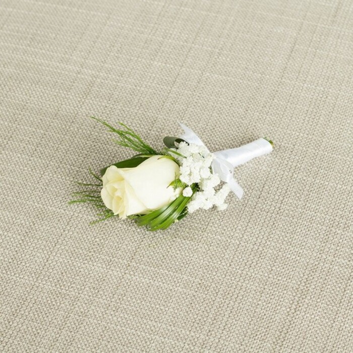 A refined white rose boutonniere lies diagonally across a softly textured, beige fabric surface, whose subtle woven pattern calls to mind the natural linens often used at wedding venues around Summertown and central Oxford. At the centre of the arrangement is a single rosebud in shades of creamy white and soft ivory, its petals tightly layered and only just beginning to open, creating a graceful, tapered form that feels both youthful and timeless. The gentle lighting in the photograph catches the outermost petals, revealing a faint sheen and delicate shading from warm cream at the base to cooler white at the tips. Nestled to one side and slightly behind the rose, fine clusters of white baby's breath form an airy frame, each tiny blossom like a dot of soft, frosted lace that adds texture without overwhelming the calm simplicity of the design. Slender, dark green leaves and a few more feathery, fern-like fronds extend from behind the rose, their shapes overlapping and curving naturally, offering contrast and depth against both the rose and the neutral background. All the stems are bound together in a smooth, pristine white satin ribbon, carefully wrapped so that no rough edges are visible, and finished with a small, well-formed bow positioned neatly beneath the bloom. The bow's loops and trailing ends lie flat, indicating thoughtful craftsmanship by an experienced florist who understands how a boutonniere needs to sit comfortably on a lapel during a full day of celebrations-perhaps from a ceremony in an Oxford college chapel through to an evening reception near the river. The overall impression is one of intimate elegance and quiet romance, suitable for grooms, best men, or cherished guests, and perfectly in keeping with the understated sophistication often sought by couples marrying in and around Oxford.