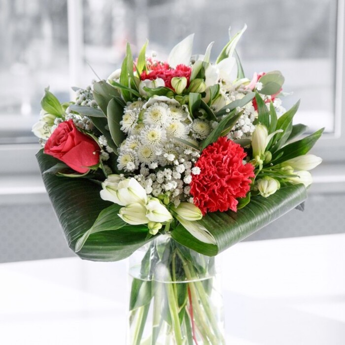 This image shows an elegant red and white bouquet arranged in a cylindrical glass vase, the stems carefully cut to an even length and immersed in clear water that speaks of freshness and skilled preparation. The focal point is a cluster of deep red roses, their velvety petals beginning to spiral open in classic romantic fashion, complemented by a richly ruffled crimson carnation at the front whose densely packed petals create movement and visual interest. Around these passionate red tones, a selection of white and cream flowers brings balance: white alstroemeria with slim, arching petals and delicate inner speckles, along with several tightly furled white lily buds poised to bloom over the coming days. Tiny white gypsophila blooms are woven throughout like a fine mist, adding lightness and a sense of celebration. Broad, glossy, dark green tropical leaves are artfully curled and wrapped around the base of the bouquet, forming an elegant green frame, while additional foliage in eucalyptus-like greens and pointed leaves gives the design depth and a natural, garden-like feel. The bouquet is placed on a pristine white surface, perhaps a dining table in an Oxford home near Summertown, with a bright, softly blurred background that suggests daylight streaming through a nearby window. The even illumination enhances the rich reds, pure whites, and layered greens without harsh shadows, creating a calm yet uplifting atmosphere. As a piece, it feels like a modern take on timeless romance, suitable as a central display for anniversaries, thoughtful birthday surprises, or a refined gift to send warm wishes anywhere in Oxford. The overall impression is one of care, intention, and heartfelt elegance, as if crafted by a florist who knows how flowers can transform a simple room into a moment.