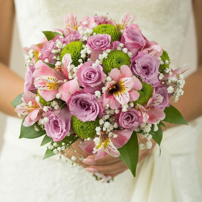 This breathtaking bridal bouquet, lovingly crafted for a wedding in Oxford's historic centre, is a harmonious blend of classical charm and fresh, modern notes. The close-up presentation reveals a lush, spherical arrangement made with care by Flowers Oxford, whose skilled florists draw inspiration from local parks like Christ Church Meadow and the tranquil Oxfordshire countryside. At the bouquet's core, soft lavender and blush pink roses blossom in various stages, each petal subtly adorned with the shimmer of morning dew. Vivid green button chrysanthemums intersperse the roses, their round, pom-pom shapes adding movement and a refreshing twist to the pastel palette. The addition of delicate pink alstroemeria, also known as Peruvian lilies, introduces intricate details-maroon-speckled centers glowing softly in the filtered light. Wispy white baby's breath, or gypsophila, fills the arrangement with a gentle airiness, echoing the pavilion-like freedom of Oxford's many open spaces. Dark green leaves nestle the design and provide a rich backdrop that accentuates the bouquet's colours and lush depth. The bride's hands, adorned with a simple band, tenderly support the stems, captured against the gown's flourished lace-a texture reminiscent of generations-old traditions in Oxford ceremonies. Altogether, this bouquet is an embodiment of romance, artistry, and celebration, perfect not only for the most elegant city weddings but also for those seeking inspiration from Oxford's timeless beauty and sophisticated floral offerings.