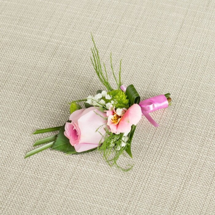 Diagonally laid on creamy beige linen reminiscent of Oxford's heritage townhouses, this floral boutonniere is a study in refined artistry and joyful celebration. The centrepiece is an immaculate blush pink rose, its petals meticulously wound into a spiralling bud, symbolising both innocence and untold stories. Flanking the rose is a feather-soft bloom-likely Alstroemeria-with petals in shades of light peach and gentle cream, adorned with subtle speckles of yellow and deep pink reminiscent of Christ Church meadows awash with wildflowers. Wisps of white Baby's Breath thread through the arrangement like morning fog drifting by Magdalen Bridge, while bright green, mounded mini mums introduce playful, rounded accents of vibrancy. Lush greens encircle the florals: upright blades suggest the neat lawns of Oxford's quadrangles and broader, deeply coloured leaves are expertly folded to anchor the base, their polished surface gleaming gently under the even daylight. A broad dark green leaf wraps precisely around the bundled stems, seamlessly integrating structure with organic flow. The finishing touch is a perfectly tied fuchsia pink satin ribbon bow at the base, its gleaming sheen creating a festive note akin to the ceremonial sashes worn at graduations near Radcliffe Square. The intricate composition, illuminated by soft, diffused light, brings together touchable textures and elegant colour harmony-crafted for the romantic moments and scholarly settings of Oxford, perfect for pinning on the lapel at a wedding, prom, or special academic celebration.