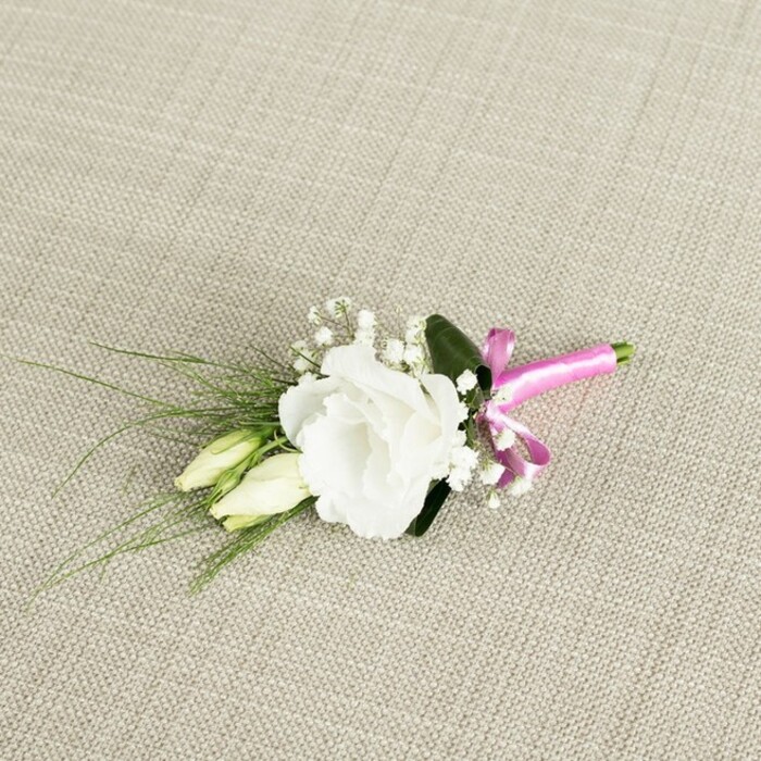 A refined floral boutonnière, ideal for a groom or groomsman, is displayed on a warm, light beige, woven fabric that resembles natural linen, its subtle weave adding a rustic yet sophisticated backdrop often seen in Oxford barn or college weddings. At the centre of the design is a fully open white eustoma, or lisianthus, its petals gently ruffled and layered in soft concentric curves that create a plush, rounded blossom with an almost cloud-like softness. Just below and to the left, two closed lisianthus buds in a creamy, pale green shade rest alongside the main bloom, their untouched tips evoking the quiet calm of a wedding morning before guests gather in places like Oxford Town Hall or the cloisters of a nearby college. Interspersed among these focal flowers are delicate clusters of baby's breath, with tiny white blossoms that give the arrangement an airy, lace-like quality and a sense of lightness. Lush greenery frames the flowers: slender, grass-like strands of dark green foliage splay gracefully towards the left, adding movement and a slightly wild, garden-inspired feel, while a broader, deeper green leaf sits discreetly behind the central bloom, offering structure and contrast. At the bottom right, the stems are carefully bound in a vivid fuchsia pink satin ribbon, its surface glossy and smooth, tied into a small yet precise bow that injects a fun, modern twist into the classic white-and-green palette. Soft, even lighting gently illuminates the petals, foliage, and ribbon without harsh shadows, emphasizing the freshness of the flowers as if they have just been prepared by Flowers Oxford for same-day delivery to a ceremony anywhere across the city, from Jericho's terraces to the leafy avenues of North Oxford, capturing a blend of timeless elegance and contemporary charm.