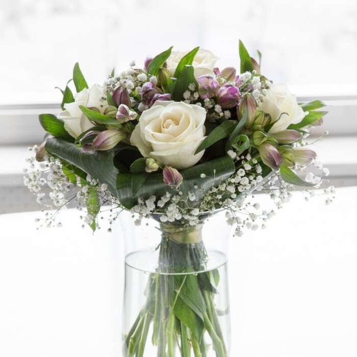 A refined bouquet of creamy white roses, lavender-tinted alstroemeria, and cloud-light baby's breath stands poised in a clear cylindrical glass vase, bathed in gentle natural light that suggests a bright but softly diffused Oxford afternoon. The roses are fully open and impeccably shaped, their velvety, ivory petals layering outward in loose spirals that create a plush, cushion-like effect at the heart of the design. Nestled around and between them, alstroemeria blooms and buds in soft lavender and muted purple provide a graceful contrast; some petals are already stretching wide, revealing deeper purple veining, while others remain in slender, tapering buds that hint at the arrangement's evolving beauty over the next few days. Filigree sprigs of white gypsophila drift among these larger blooms, filling small gaps so that the bouquet feels full and balanced while adding a light, lacy texture that keeps the overall look airy. A collar of rich green foliage-from broad, dark leaves to finer, more flexible stems-encircles and supports the flowers, shaping the bouquet into a rounded, symmetrical dome. Through the transparent glass vase, the neatly grouped stems are visible, cut cleanly and arranged to radiate naturally into the water, as a skilled florist would do in a workshop just off Cowley Road or near the Covered Market. The lighting is calm and even, smoothing any harsh shadows and allowing the soft whites and lilacs to glow with an almost ethereal luminosity. This arrangement feels perfectly suited to expressing gentle sympathy, celebrating a quiet anniversary in North Oxford, or simply brightening a hallway in Headington with a sense of purity, hope, and graceful, understated elegance.