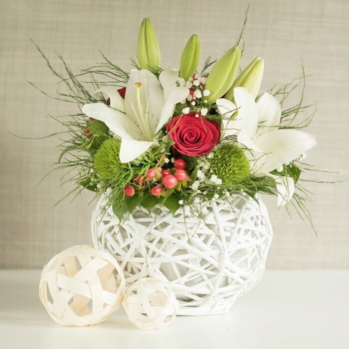 A sophisticated floral centerpiece sits quietly radiant on a smooth white tabletop, its design perfectly suited to a wedding breakfast or elegant celebration in one of Oxford's historic venues near Broad Street or St Giles'. The arrangement is cradled in a striking spherical white vase, formed from interwoven strands that create a light, basket-like texture, allowing glimpses of shadow and depth between the curved lines. Rising from the vase is a lush display of flowers and foliage, built around several generous white lilies. Some lilies are fully open, their broad, satin-like petals arching outward and curling slightly at the tips, revealing soft speckles and bold, golden stamens at the centre. Others remain as tightly closed green buds, pointing gracefully upward and hinting at fresh blooms in days to come, a symbolic gesture of new beginnings that suits wedding celebrations. Within this sea of white, a single red rose stands out, its velvety petals wrapped in a near-perfect spiral, adding a deep note of romance and drama among the lilies. Two or more bright green pompon-style flowers, probably chrysanthemums or green dianthus, nestle near the base of the arrangement, their rounded, tufted heads lending a playful, tactile contrast to the smooth lily petals. Tiny white gypsophila clusters lace through the bouquet like mist, while small red berries bring a subtle pop of warm colour, slightly reminiscent of winter hedgerows around Port Meadow. Layered greenery fills out the design with feathery fronds and glossy leaves, cascading softly over the vase's edge. To the left, two decorative woven spheres in pale, natural tones echo the vase's texture and balance the composition. Set against a softly textured neutral backdrop, this arrangement radiates calm elegance, ideal for wedding table décor or refined events across Oxford.