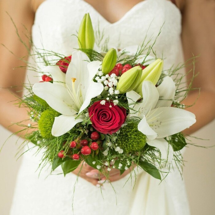 Captured in a luminous close-up, a bride in a strapless white wedding gown gently clasps an exquisite bouquet that radiates both tradition and personal expression. The upper bodice of her dress-subtly patterned and crafted from fine Oxford textiles-serves as the perfect canvas for the artistry blooming in her hands. Several large, pure white oriental lilies command the bouquet, some in full blossom with glistening golden stamens visible, others still sealed in pale green buds, their fresh promise echoing the renewal seen every spring in Oxford's Botanic Garden. Rich crimson roses anchor the centre, each tightly curled petal emblematic of enduring love and centuries-old romance found among the city's cobbled lanes. The ensemble is softened by delicate mists of white baby's breath, while striking red hypericum berries and whimsical orbs of green-resembling mossy trick dianthus-add joyful contrast and textural intrigue. Wispy ferns and broad, glossy foliage intertwine below, creating a lush, slightly wild bouquet reminiscent of Oxford's riverside meadows in full flourish. Perfect for gracing a city wedding from Magdalen College to Jericho, this bouquet feels personal and timeless-a vivid testament to the thoughtful work of local florists and a celebration of love in the heart of Oxford.