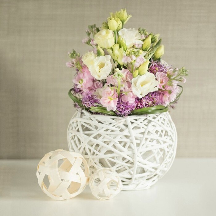 A carefully styled floral still life unfolds on a bright, white surface, where a rounded, open-weave white vase takes centre stage, overflowing with soft, romantic blooms. The vase itself resembles a delicate sphere woven from white wicker or painted rattan, its airy latticework creating a sense of lightness and movement, almost like a modern art piece you might spot in an independent gallery off Oxford's Cowley Road. Emerging from this sculptural base, a generous bouquet of eustoma, or lisianthus, in creamy white and very pale yellow fills the upper portion of the arrangement. Their petals are finely ruffled and layered, giving a plush, petal-soft texture that suggests quiet luxury and gentle movement. Interspersed among them are slender, upright stems of stock in shades of soft pink and lavender, whose vertical lines lend structure and height, guiding the eye upward in a graceful sweep. At the lower level of the bouquet, small, deep purple filler blooms cluster together, creating a rich, textural foundation that emphasises the pastel tones above, while a close-fitting ring of deep green foliage encircles the design, providing a crisp, botanical frame. To the left of the main arrangement, two decorative spheres in light natural wood or rattan rest casually on the tabletop, their open basket-weave pattern echoing the vase and reinforcing the natural, handcrafted theme. Behind, a warm beige or greige wall with a subtle vertical texture forms a soft, neutral backdrop, allowing the colours and shapes of the flowers to stand out without distraction. The gentle, diffused lighting and harmonious palette evoke feelings of calm and understated elegance, making this arrangement perfect for an intimate dinner, a refined birthday surprise, or a welcoming reception display in an Oxford office or townhouse.