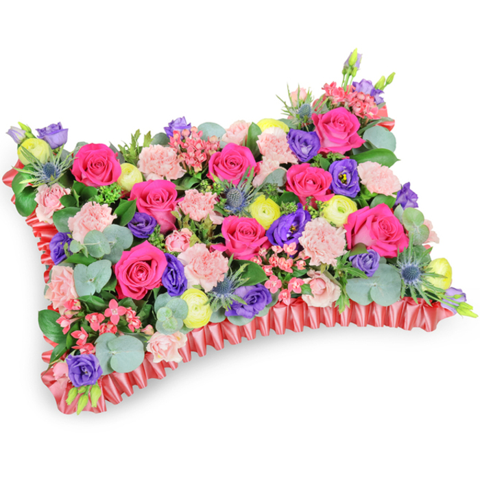 This carefully staged photograph shows a pillow-style funeral tribute photographed from above and slightly to one side, resting on a stark white background that highlights every hue and texture. The surface is a dense quilt of hot pink roses-some with the velvet sheen of newly opened petals, others still tight and hopeful-balanced by pale pink carnations that lend a delicate, frilled texture throughout. Bright, buttery-yellow ranunculus provide sunny, uplifting accents among the pinks, while sumptuous purple lisianthus contributes cool, bell-shaped depth. The arrangement is given a touch of structural drama by several silvery-blue Eryngium thistles; their spiky, almost architectural presence injects a contemplative contrast against the softer blooms. Eucalyptus leaves, round and silver-tinged, thread through the composition alongside darker green foliage, offering both scent and a natural backdrop that visually cushions each bloom. The whole piece is edged in a ruffled, pleated deep-red satin ribbon which crisply defines the rectangular pillow shape and adds a subtle sheen at the margins. Under soft, diffused lighting the textures read clearly-velvet, ruffle, spike and leaf-evoking a tactile sympathy and a quiet celebration of memory. Designed for memorials and funerals across Oxford, whether laid at services near the University Church or placed at gravesides by Port Meadow, this floral pillow speaks of remembrance, warmth and a carefully tended affection.