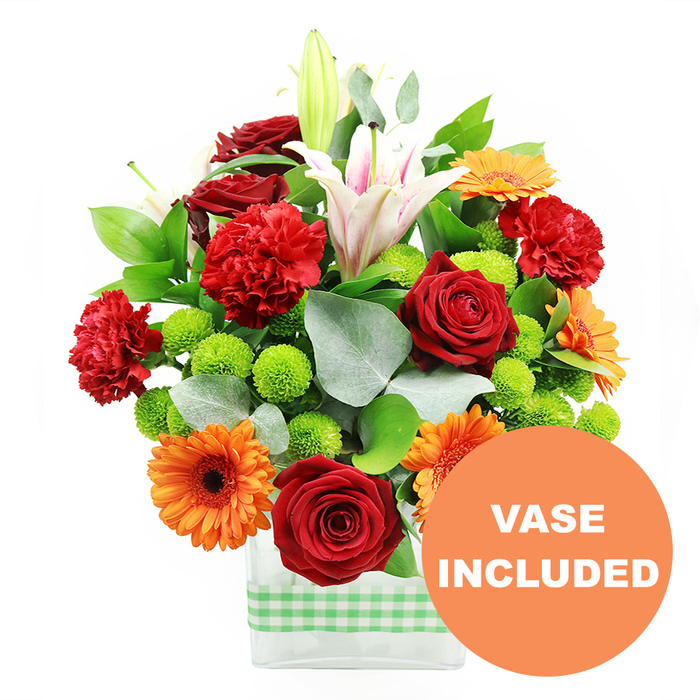 A lush, colour-packed bouquet stands proudly in a sleek rectangular glass vase, captured against a clean, white backdrop that lets every hue and texture stand out clearly, much like a bright arrangement in an Oxford café window. The vase is wrapped with a band of light green and white gingham ribbon around its lower half, lending a touch of rustic charm that feels at home in neighbourhoods from Summertown to Headington. Emerging from this base is a dense, thoughtfully layered assortment of fresh flowers. Deep crimson red roses with tightly furled, velvety petals anchor the arrangement, joined by generous clusters of red-orange ruffled carnations that add volume and a soft, cloud-like texture. Intermingled with these are vivid orange gerbera daisies, each bloom perfectly flat with a strong dark centre, their petals forming radiant circles that bring a sense of joy and cheer. Tall white Oriental lilies rise gracefully above the rest, some fully open to reveal pale pink stripes along the inner petals and delicate stamens dusted with golden pollen, while others remain sealed in elegant buds, hinting at continued unfolding beauty. Nestled between the larger blossoms, bright lime green pompom chrysanthemums appear as tidy, spherical buttons, adding a playful, almost contemporary graphic feel. The arrangement is framed and filled with rich green foliage and the cooler tones of silver-green eucalyptus leaves, which spill and curve naturally around the edges, giving movement and depth. Through the clear sides of the vase, the green stems can be seen crossing in the water. In the bottom right corner of the image, a bold circular orange label with white capital lettering clearly states VASE INCLUDED, signalling that this design is delivered as shown, ready to brighten living rooms, student flats, or offices throughout Oxford.