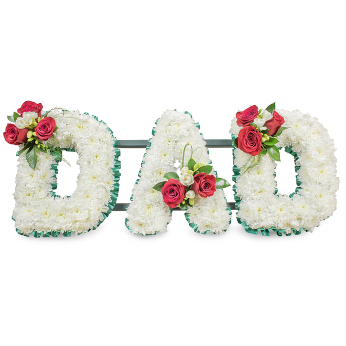 A carefully composed funeral tribute spells the word 'DAD' in large, three-dimensional letters, each densely covered with small, immaculate white chrysanthemum blooms that form a uniform, velvety surface. The white chrysanthemums read as a symbol of purity and steadfast devotion; their compact heads create soft sculptural planes that are subtly highlighted by gentle, even lighting. Each letter is outlined in a deep emerald-green pleated ribbon or ribbed foliage trim, which provides a crisp, refined border and helps the letters hold their classic block form. On the upper left curve of the first 'D', within the negative space of the 'A', and on the upper right curve of the final 'D', matching clusters of rich red roses and delicate white freesia have been artfully arranged. These clusters introduce a striking colour counterpoint: the velvety red petals of the roses convey warmth and respect, while the freesia buds add a hint of perfume and a lighter, airy texture. Sprays of lush dark green foliage and subtle loops of fine greenery thread through each cluster, creating movement and framing the focal blooms. Thin dark metal or painted green support bars sit behind the letters, mostly concealed but essential for stability when the piece is placed at a service table or beside a graveside. Set against a bright white backdrop, the tribute's contrasts, textures and restrained palette speak to formal remembrance and personal affection - a design often selected by families across central Oxford, near Broad Street or the Botanic Garden, seeking a dignified, heartfelt way to honour a father.