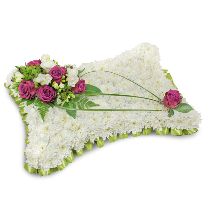 A traditional rectangular floral pillow, expertly assembled to symbolize peace and final rest, occupies a crisp white studio space with a faint shadow beneath that highlights its pillowy profile. The entire cushion is densely carpeted with small, immaculate white chrysanthemum florets whose compact, rounded petals create a tactile, cloudlike surface that reads as tender and reverent. Anchoring the composition in the upper-left is a vibrant focal cluster of deep fuchsia roses-each velvety bloom unfurling with a plush, comforting presence-interspersed with slender white freesia and occasional creamy lisianthus that lift the colour and add a subtle, fragrant note. Verdant fern fronds and bright green foliage provide a lush backdrop for the accent blooms, giving the grouping a natural, garden-fresh depth. Three slender stems of dark green bear grass arch gracefully from the cluster across the pillow, forming elegant lines that guide the gaze toward the bottom-right where a solitary fuchsia rose nests among leaves, mirroring the principal spray. The edge of the pillow is finished with a folded lime-green satin ribbon that creates a scalloped, ruffled border, introducing a soft contrast and a handcrafted polish. The overall effect is one of calm dignity and thoughtful craftsmanship-an arrangement suitable for honouring a life at a funeral near the Botanic Garden or a memorial service in Jericho. Prepared by seasoned florists at Flowers Oxford, this piece communicates sympathy with quiet, enduring beauty and is available for local delivery within Oxford.