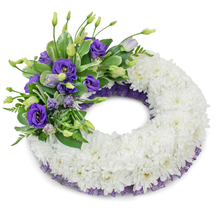 A graceful circular funeral wreath photographed on a pure white background, composed to offer silence and solace through floral form. The body of the wreath is a thick, uninterrupted ring of immaculate white chrysanthemums - cushion varieties whose abundant, ruffled petals are densely massed to create a soft, pillowed texture that suggests peace and continuity. At the upper-left section an abundant accent of purple lisianthus provides a focal point: fully opened deep-violet blooms with delicate yellow centres sit beside paler lavender buds and upright stems, their silky petals introducing a gentle contrast in hue and surface. Sprigs of fresh green foliage - glossy leaves with subtle variegation alongside airy, fern-like fronds - weave through the purple cluster, adding depth, movement and a restrained green scent reminiscent of the trees along High Street. Peeking out beneath the main white body is a fine trim of dark purple mesh or lace along both inner and outer edges, a subtle flourish that echoes the lisianthus tones and lends refined definition to the wreath's silhouette. A soft, diffused shadow underlines the arrangement against the white field, emphasizing the wreath's rounded volume and the meticulous craftsmanship of each flower placement. Suitable for memorial services at local churches, crematoria, or a quiet farewell near Magdalen Bridge or Summertown, the composition communicates sympathy, respect and a careful attention to colour and proportion.