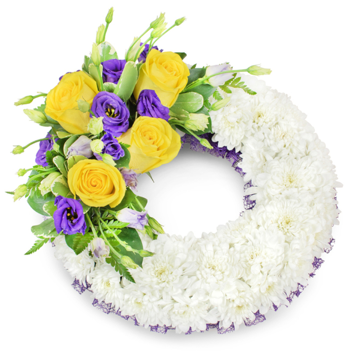 Presented against a clean white backdrop, this circular funeral wreath is a study in respectful contrast and careful craftsmanship. The principal body of the wreath is formed from a dense carpet of pristine white spray chrysanthemums - petals layered so uniformly they create a serene, textural plane that suggests purity and repose. On the upper left, a focused spray introduces colour and narrative: fully opened yellow roses in tones of butter and gold, their scent quietly suggested, sit beside clusters of deep purple lisianthus (Eustoma) whose elegant, cup-shaped blooms offer a stately softness. Small, paler lisianthus buds and delicate green stems peek through, adding dimensionality and the fresh breath of garden growth. A variety of greens - glossy leaves, subtly variegated foliage and fine fern fronds - frames the flowers, providing a verdant backdrop that enhances the vividness of the roses and lisianthus. Around the inner and outer edges of the white base a slender dark purple mesh ribbon is just visible, a refined trim that ties the whole palette together without overwhelming the wreath's quiet dignity. This piece is composed with the kind of clarity suited to memorials and grave-side services across Oxford - from tributes placed at St Giles to floral remembrances delivered within Summertown - offering a composed, heartfelt expression of condolence that balances purity, warmth and solemn beauty.