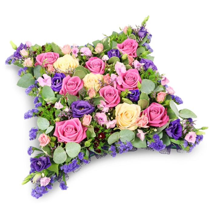 The photograph presents an elaborate floral cushion tribute, positioned centrally against a crisp white background that casts only a faint, soft-edged shadow beneath it. The arrangement is densely constructed, forming a gentle, pillow-like rectangle whose rounded sides suggest comfort, rest, and quiet support. Dominating the surface are generous roses in a carefully balanced palette: bold fuchsia blooms draw the eye, while softer pastel pink and buttery cream roses offer a tender contrast, some fully opened to reveal their swirling hearts, others still slightly cupped as if holding unspoken thoughts. Interspersed among these are deep violet-purple lisianthus, their ruffled, almost silk-like petals adding a touch of drama and serene depth, like the rich tones of an evening sky over Oxford's spires. Delicate clusters of tiny lavender and lilac filler flowers, resembling statice or limonium, fill every small gap, ensuring the cushion feels lush and complete with no bare patches. Embracing the flowers is a collar of mixed greenery: rounded, silvery eucalyptus leaves with their soft, powdery tone, partnered with slender, ferny foliage that adds a darker, feathery outline. Together, they lend hints of cool woodland fragrance and textural contrast that you might associate with a stroll through University Parks or along the towpath by the river. The overall impression is one of dignified beauty and heartfelt remembrance, a design often chosen by families in Oxford to rest on a coffin, mark a grave, or accompany a memorial service, expressing sympathy, love, and the wish for gentle, eternal peace.