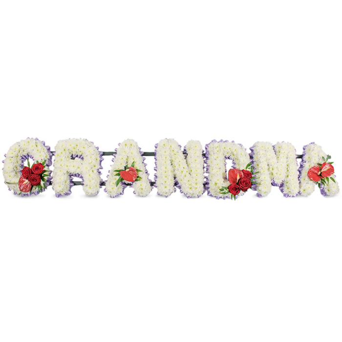 This photograph captures a dignified floral tribute spelling out the word GRANDMA in bold capital letters, designed as a loving farewell for a cherished grandmother, perhaps destined for a chapel of rest or cemetery near Oxford's tranquil green spaces. Each individual letter is composed of closely packed white cushion chrysanthemums, the uniform, button-like blooms creating a velvety, cushiony texture that appears soft to the touch. The flowers are arranged so tightly that no gaps are visible, forming crisp letter shapes that can be easily read from a distance. Around the edge of every letter runs a faint trim of pale purple ribbon or backing, which peeks out delicately between the petals and the framework, giving the tribute a gentle lavender halo that softens the strong outlines and adds depth. Rich red accents, symbolising love and remembrance, are placed at key points along the word. On the letter G, a small spray of deep red roses is arranged alongside a distinctive variegated red-and-white calla lily that tilts gracefully outward. The first A is marked by a single heart-shaped red anthurium, its smooth, glossy surface contrasting with the feathery chrysanthemums. In the middle, the M carries another cluster of red roses, enhanced by greenery and a matching red anthurium that echoes the earlier motif. Finally, the last A features its own red anthurium, completing the line of heartfelt red highlights. The letters rest on a discreet, dark green stand or rail that supports them in a neat horizontal row. Photographed against a clean white background, the arrangement appears bright and composed, reflecting both craftsmanship and compassion, much like the thoughtful funeral floristry available throughout Oxford's High Street, Jericho, and Summertown.