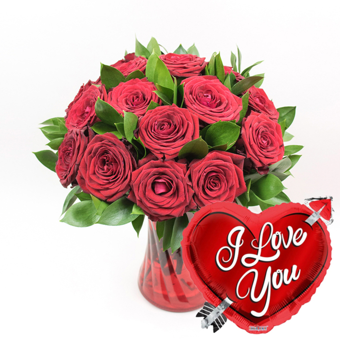 This image captures a dramatic, love-soaked still life: a ruby-red glass vase, tall and gently fluted, overflowing with an abundant cluster of deep crimson roses. The blooms are lush and layered, with velvety petals that spiral outward in tight curls at the centre and soften into broader, more relaxed edges at the tips. Some roses appear fully open, proudly displaying their hearts, while others stay slightly cupped, suggesting a love still unfolding. Threaded through the arrangement, rich green foliage with a glossy sheen creates a vivid contrast, giving structure and framing the flowers so they seem to rise like a crimson fountain. The vase itself, tinted a warm ruby hue, has vertical ridges that catch the light and mirror the length of the stems inside, grounding the arrangement with both colour and texture. The entire bouquet stands against a pristine white background, making the reds and greens leap out as clearly as the college spires do against an Oxford sky. In the lower right foreground, partially overlapping the vase, sits a gleaming, metallic red heart-shaped foil balloon. Its reflective surface shows gentle highlights and shadows, and across its centre flows the message I Love You in crisp, white cursive script. A shining silver Cupid's arrow appears to pierce the heart from lower left to upper right, adding a playful yet sincere symbol of devotion. The composition conveys the feeling of a carefully planned romantic gesture, perfect for a surprise delivery to a loved one in Cowley, a special anniversary dinner in central Oxford, or any moment when a bold, unmistakable declaration of love is called for.