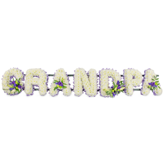 The image shows a moving and carefully composed GRANDPA funeral tribute arranged in flowers, the word set out in big, bold capital letters across a pure white background. Each letter is completely covered with compact, snowy-white chrysanthemums or carnations, the blooms arranged so closely that they form a continuous, pillow-soft surface with no visible stems or gaps. The white petals create a gentle, almost glowing effect, reminiscent of the quiet light that falls across the cloisters and college chapels of Oxford. Along the contours of the letters, thoughtful bursts of deep purple and soft lavender flowers are woven into the design, paired with fresh green foliage. These accents are nestled within the curve of the 'G', in the open triangles of both 'A's, along the upright of the 'D', and around the rounded bowl of the 'P', bringing subtle definition and a sense of warmth to the tribute without overwhelming its calm, white base. The purple tones, perhaps lisianthus or other dainty funeral blooms, symbolise dignity and reflection, while the greenery adds a natural, living contrast. Beneath the line of letters, a sturdy dark green frame runs horizontally, mostly hidden but vital for supporting the arrangement on an easel, hearse, or graveside stand, whether delivered to a church service off the Cowley Road or a quiet ceremony in one of Oxford's village churches. The overall impression is one of deep respect and enduring love, a personalised floral message that allows family to honour their grandfather by name, creating a focal point for remembrance and shared stories during a time of loss.