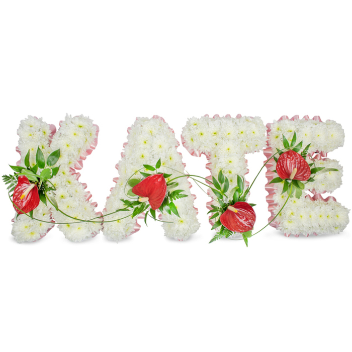 An elegant, personalised floral tribute spells KATE in three-dimensional capital letters, each letter meticulously covered with a dense carpet of small white chrysanthemums to create a uniform, plush texture that reads as peaceful and pure. A soft, pale pink ribbon with a ruffled edge follows the outer contours of each letter, its delicate colour adding definition and a tender, almost fabric-like softness. Bright red anthuriums are set into the chrysanthemum field at carefully considered points, their glossy, heart-shaped spathes and protruding spadices supplying vivid contrast and a heartfelt focal note. Lush green foliage - broad glossy leaves, slender fern fronds and fine trailing vines - is woven throughout the arrangement, framing the letters and linking them with natural curves that suggest continuity and remembrance. The letters are presented against a clean white backdrop with a low shadow beneath them, creating subtle depth and a quiet, sculptural quality. This piece is intended for memorial settings: a church service near the Covered Market, a funeral luncheon at a local hall, or a graveside gathering in the green spaces around University Parks. The overall impression is one of restrained beauty and reverence; the cool whiteness of the chrysanthemums tempered by the warmth of red anthuriums and the gentle blush of ribbon conveys sympathy without ceremony, a calm testament to a life remembered. The floral textures invite a gentle, contemplative touch, and the design demonstrates the care and technical skill of experienced funeral floristry.