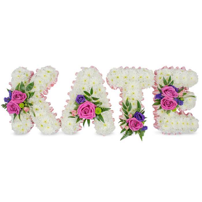 An elegant, solemn floral name tribute spelling KATE, presented as four freestanding letters made from a dense, immaculate carpet of white chrysanthemums that form smooth, pillowy faces for each character. A pale pink satin ribbon delicately traces both outer and inner contours, offering a soft edge that complements the white while introducing a gentle, compassionate tone. Each letter carries a carefully arranged cluster of accent blooms-intense fuchsia-pink roses, softer pastel pink spray roses, and small deep purple or blue accent stems, likely statice or lisianthus-selected to provide poignant contrast and visual weight without overwhelming the purity of the chrysanthemum base. Sprays of greenery, from lacy fern fronds to elegant eucalyptus leaves, cradle these clusters and introduce varied leaf textures and cool green notes that calm the palette. The floral placements are composed with thoughtfulness: a prominent grouping on the K's diagonal, a tender cluster at the lower right of the A, a grounding spray at the T's base, and two composed accents on the E's middle and lower bars-creating a measured, reverent flow across the name. Under soft daylight the white blooms read as luminous, while the pinks and purples offer compassionate color, evoking both celebration of a life and quiet mourning. This bespoke tribute is a classic choice for memorial services, graveside gatherings, or a church send-off in Oxford, familiar to families who attend services near the High Street or prefer a memorial close to Magdalen College; it speaks of careful craftsmanship and sincere sympathy.